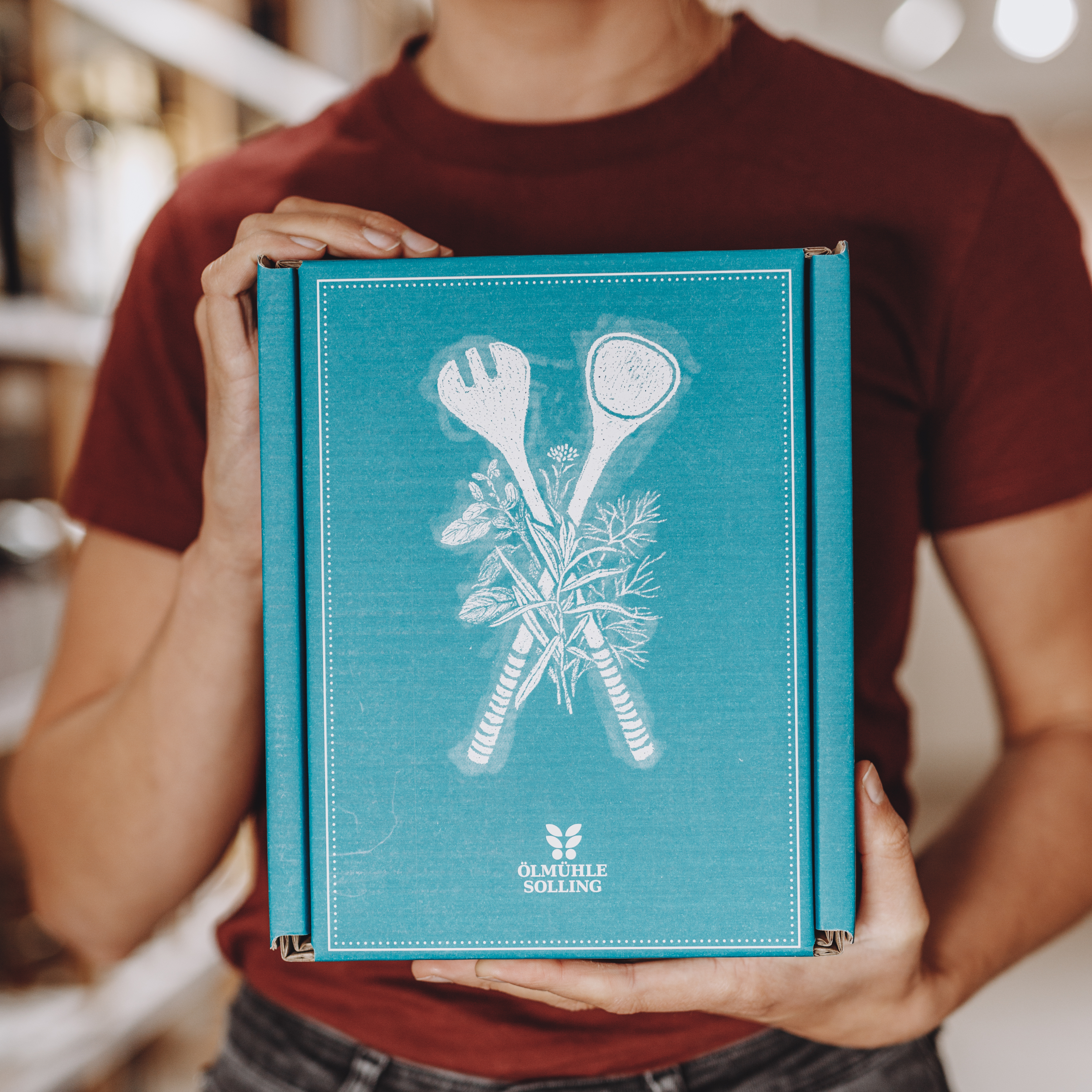 Eine Person hält ein Geschenkset der Ölmühle Solling mit der Aufschrift 'Klassische Lieblinge' in den Händen. Die Verpackung ist türkis und zeigt eine weiße Illustration von Küchenutensilien und Kräutern. Das Set enthält drei 250 ml Flaschen und wird als Delikatessenset präsentiert.