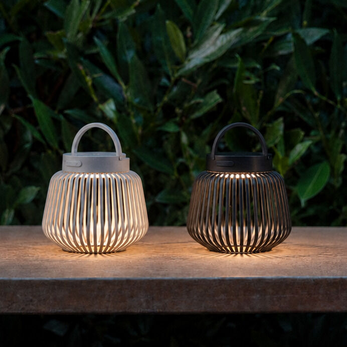 Zwei kabellose LED-Tischleuchten der Marke blomus in Rattan-Optik, eine in Weiß und eine in Granite Grey, stehen auf einer Steinplatte vor einem dichten, grünen Blattwerk und verbreiten sanftes Licht. Ideal für Drinnen und Draußen, modernes Design.