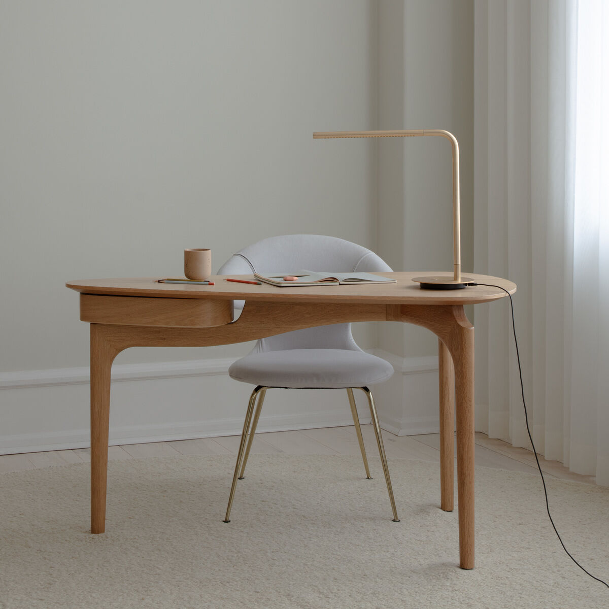 Moderner Schreibtisch aus hellem Holz mit abgerundeten Kanten steht vor einer hellen Wand und einem Fenster mit weißen Vorhängen. Auf dem Schreibtisch befinden sich eine minimalistische Lampe, ein Becher und Unterlagen. Ein gepolsterter Stuhl steht davor, alles in einer hellen, ruhigen Umgebung auf Teppichboden.