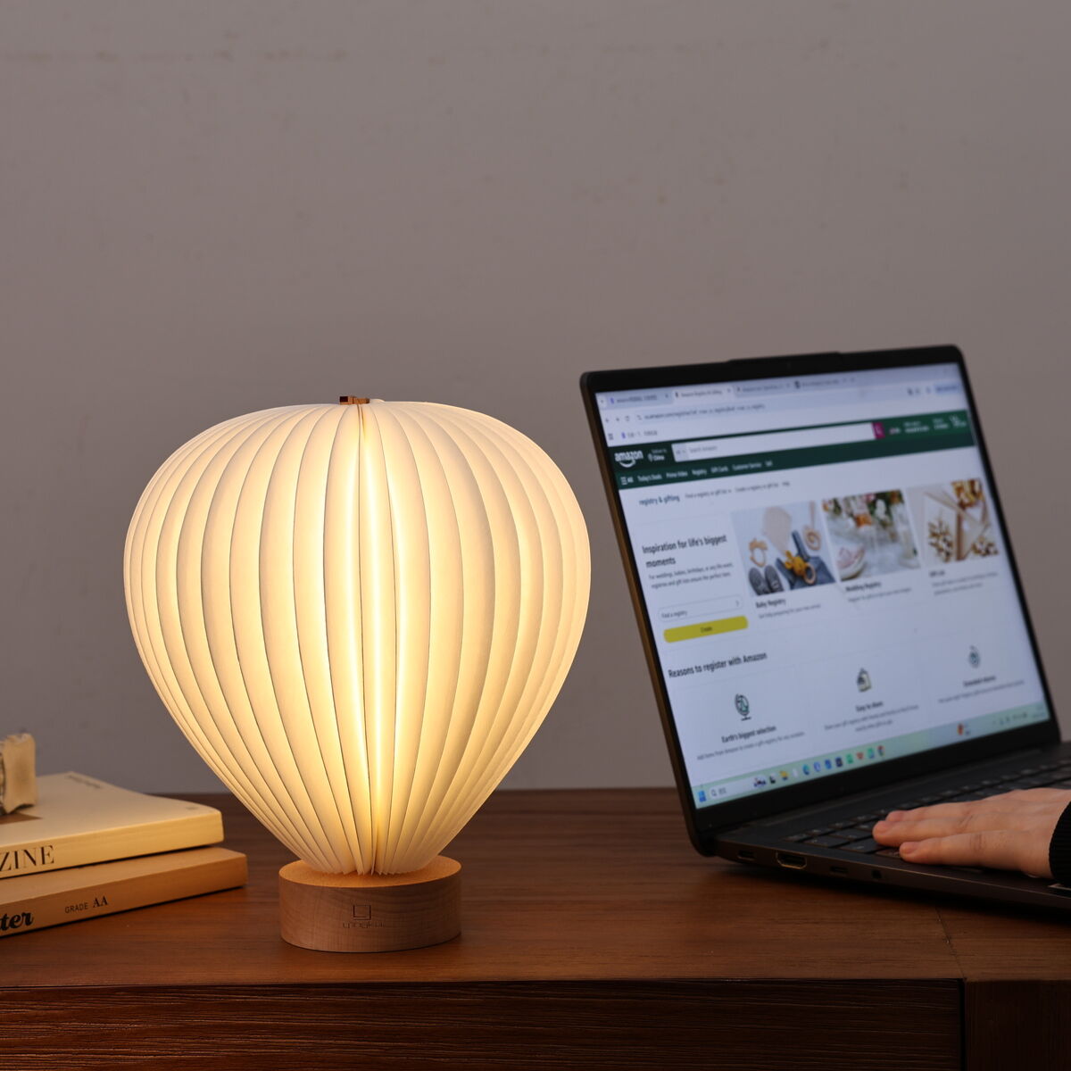Auf einem Holzschreibtisch steht die stilvolle Leuchte »Aeris Light« von Gingko Design Ltd in Form eines Heißluftballons mit einer Basis aus Eschenholz. Die Lampe strahlt ein warmes, weiches Licht aus. Daneben liegen mehrere Bücher und ein aufgeklappter Laptop, an dem eine Person arbeitet.