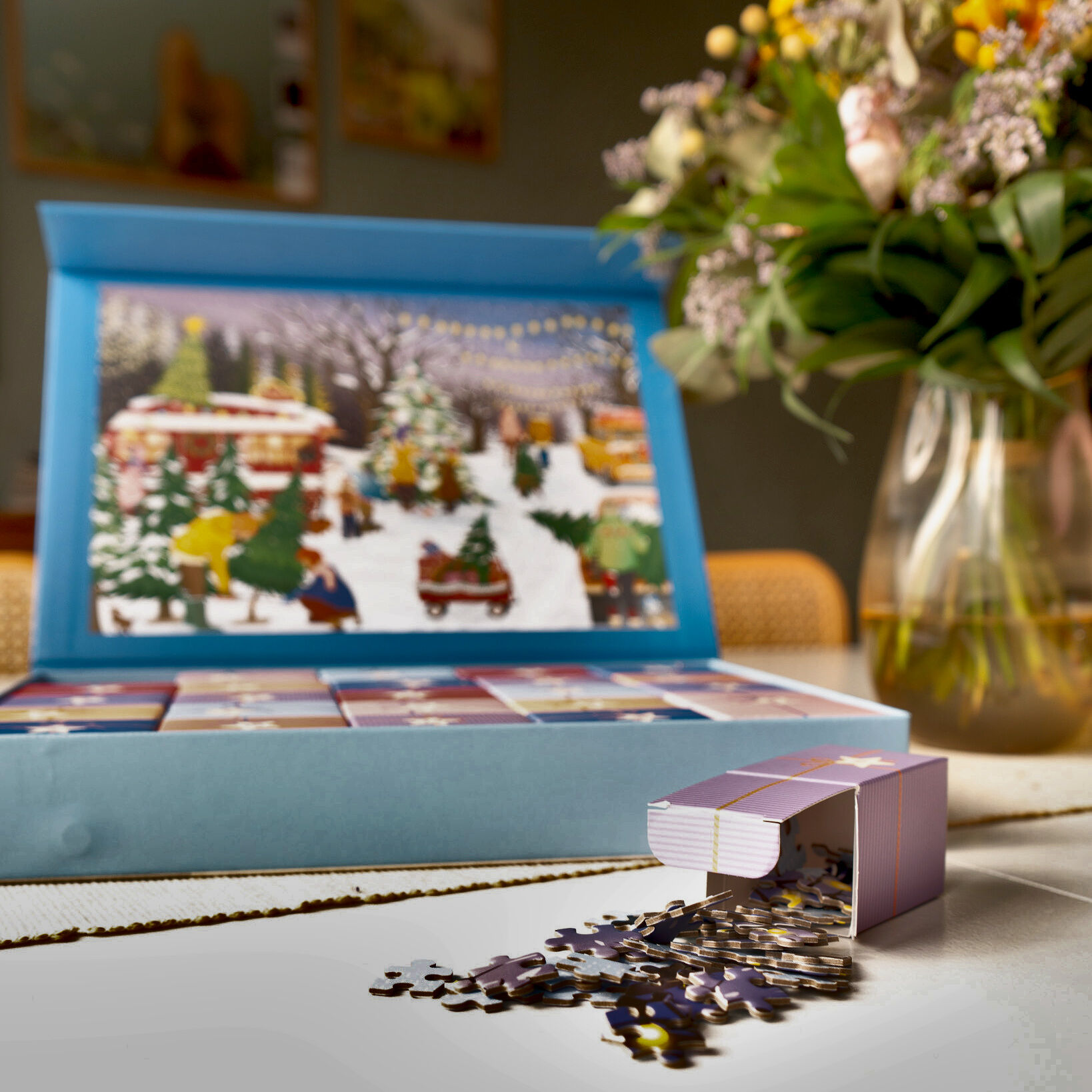 Ein Puzzle-Adventskalender mit dem Motiv von Weihnachtsbäumen und Schneeflocken steht auf einem Tisch. Im Vordergrund ist eines der geöffneten Adventsfächer zu sehen, aus dem Puzzleteile herausgleiten. Im Hintergrund ist ein winterliches Puzzlebild mit festlich geschmückten Bäumen, winterlicher Landschaft und Lichtern zu erkennen. Daneben steht eine Vase mit frischen Blumen. Die Szene vermittelt eine stimmungsvolle Adventsatmosphäre.