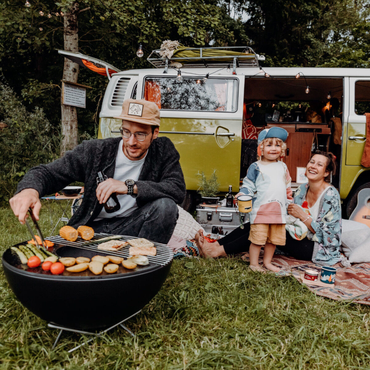 Eine Familie genießt ein Picknick draußen neben einem alten Bulli. Ein Mann grillt Fleisch und Gemüse, während eine Frau mit zwei Kindern auf einer Decke sitzt.