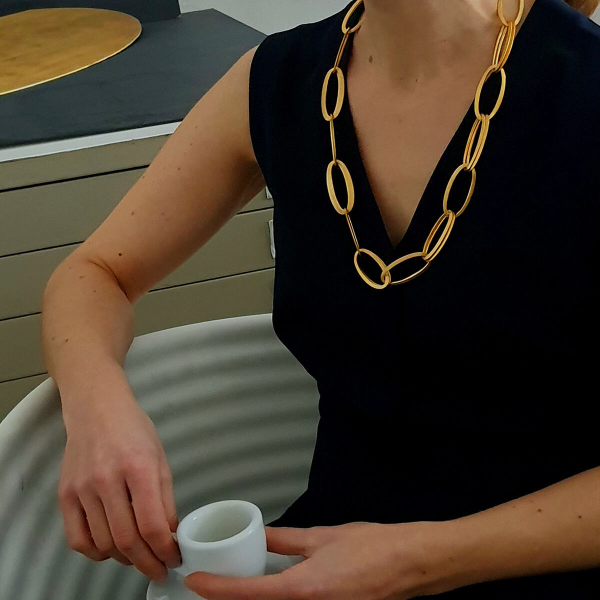 Eine Frau trägt ein feingoldplattiertes Collier »GRACE« von HML Berlin aus großen, ovalen Gliedern über einem schwarzen, ärmellosen Kleid. Sie sitzt an einem Tisch mit weißer, strukturierter Oberfläche und hält eine schlichte, weiße Tasse in der Hand.