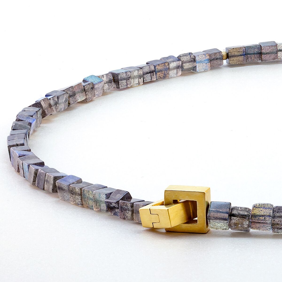 Abgebildet ist das Labradorit Collier von Menard. Die Kette besteht aus kleinen, kubischen Labradorit-Steinen mit irisierenden Grautönen, die von mattierten goldplattierten Silberkugeln unterbrochen werden. Mittelpunkt ist eine eckige, goldplattierte Patentschließe.