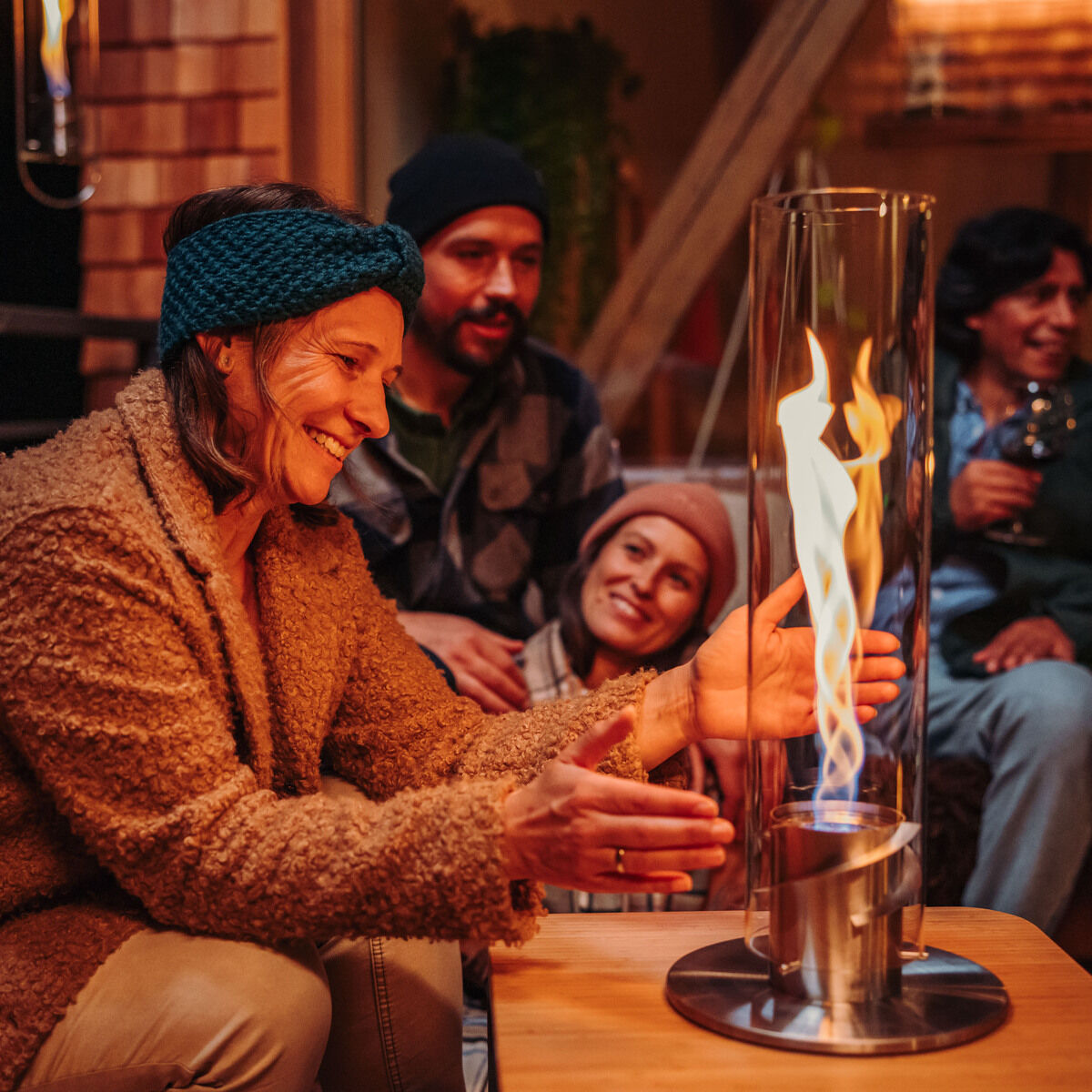 Vier Menschen sitzen in einer gemütlichen Umgebung. Eine Frau hält ihre Hände in die Nähe eines großen, zylindrischen Tischfeuer »SPIN 1200«, das auf einem Holztisch in der Mitte steht. Die Flammen sorgen für eine warme und einladende Atmosphäre.