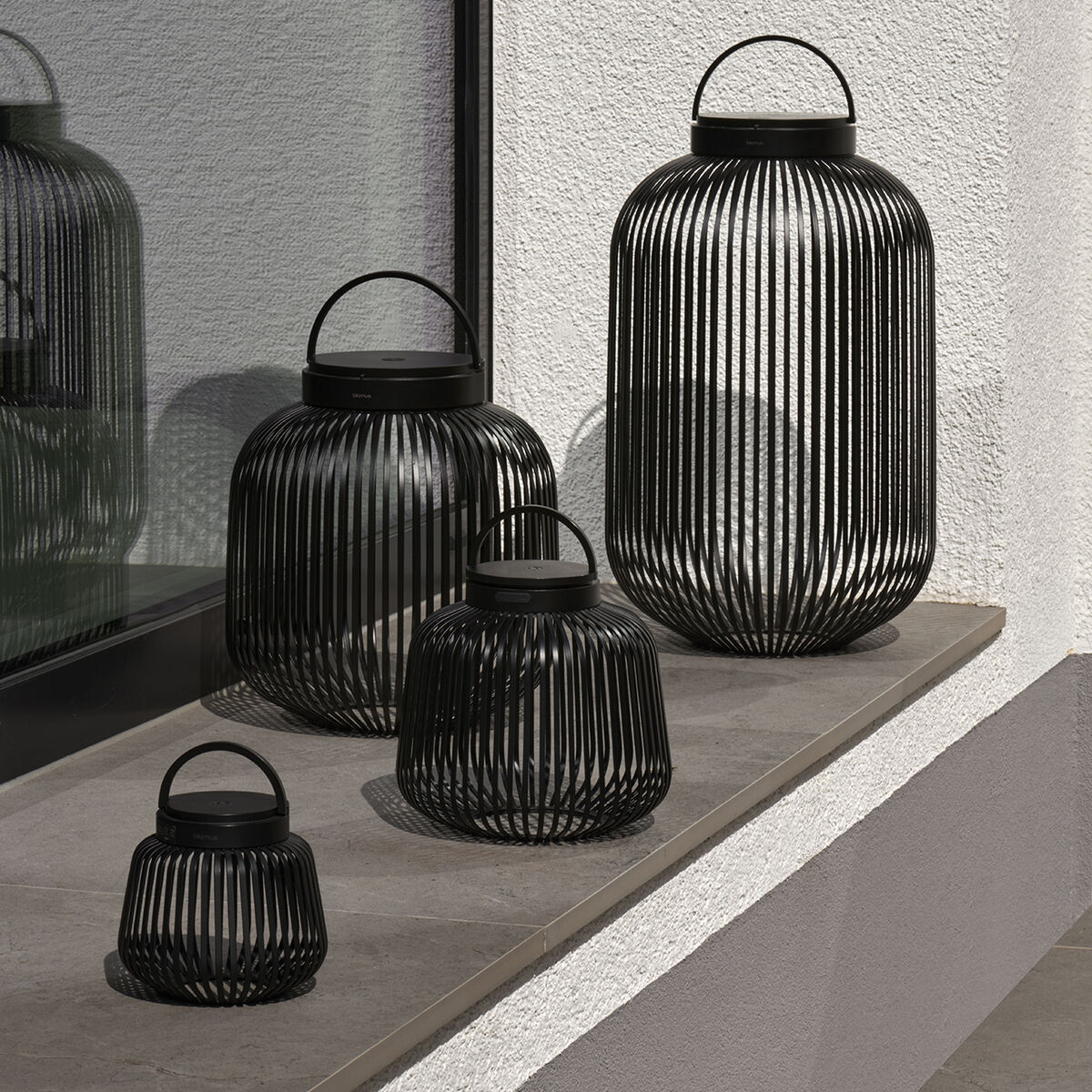 Vier schwarze LED-Leuchten 'LITO' von blomus in unterschiedlicher Größe stehen auf einer Steinbank vor einer weißen Wand mit Fenster. Die filigranen, aus Metallstäben gefertigten Lampen wirken modern und sind sowohl für den Innen- als auch Außenbereich gestaltet.