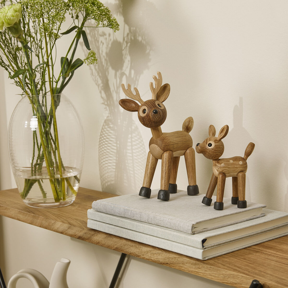 Zwei stilisierte Holzfiguren in Form eines Hirsches und eines Rehs stehen auf einem Stapel hellgrauer Bücher auf einem Holzregal. Daneben befindet sich eine Glasvase mit hellgrünen und weißen Blumen. Im Hintergrund sind eine weiße Keramikkanne und eine cremefarbene Wand sichtbar. Die Szene wirkt modern, natürlich und skandinavisch inspiriert.