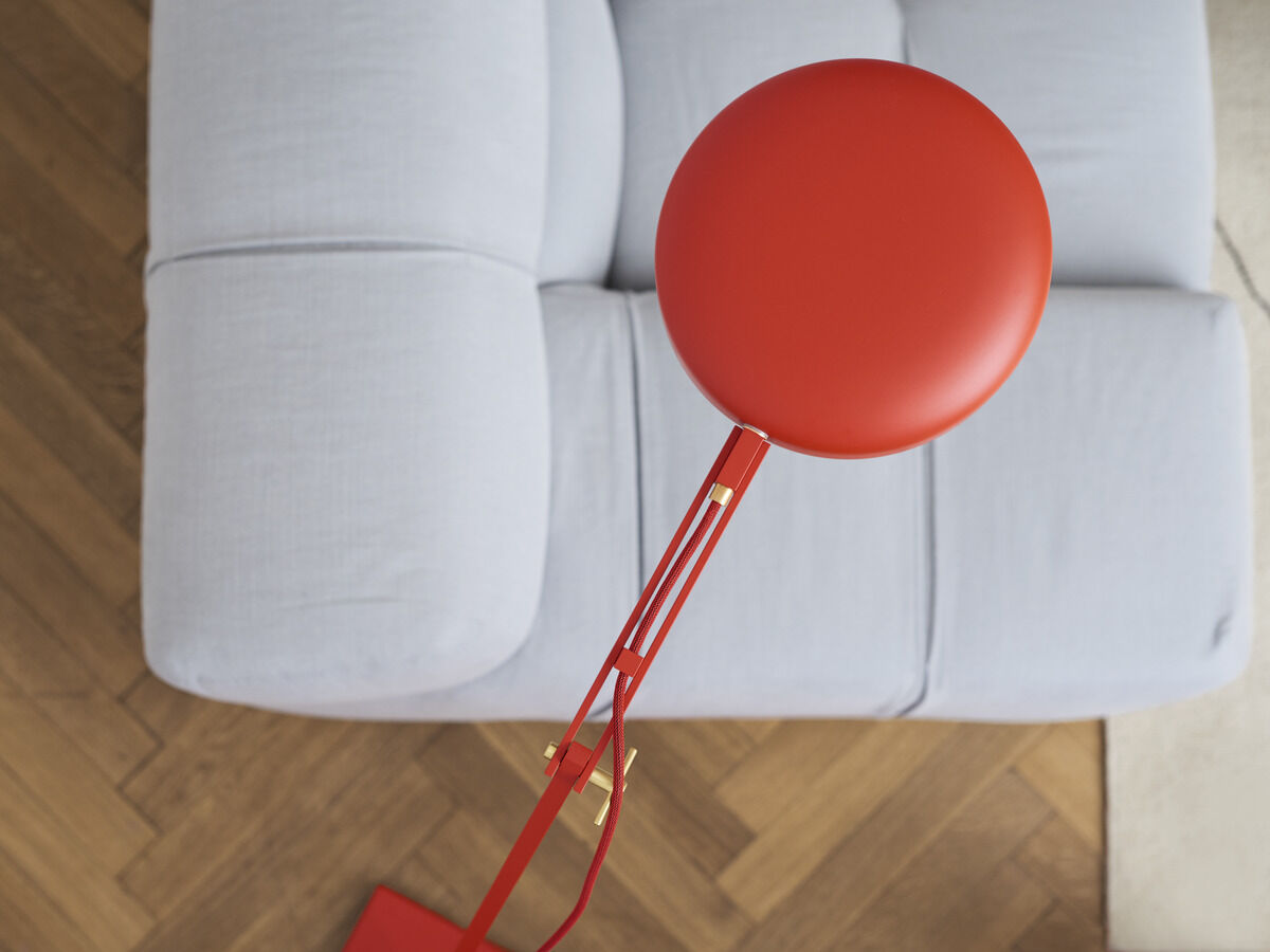 Eine rote Stehleuchte mit modernem Design steht neben einem hellgrauen Sofa auf einem hellbraunen Holzboden.