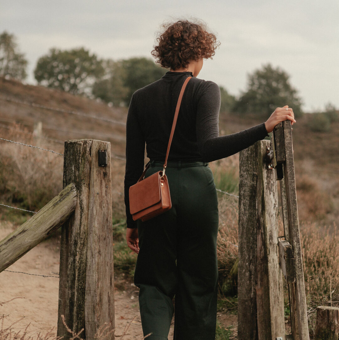 Eine Frau im schwarzen Oberteil und grünen Hosen trägt über die Schulter die braune Tasche Audrey Mini und geht draußen einen holzigen Pfad entlang.