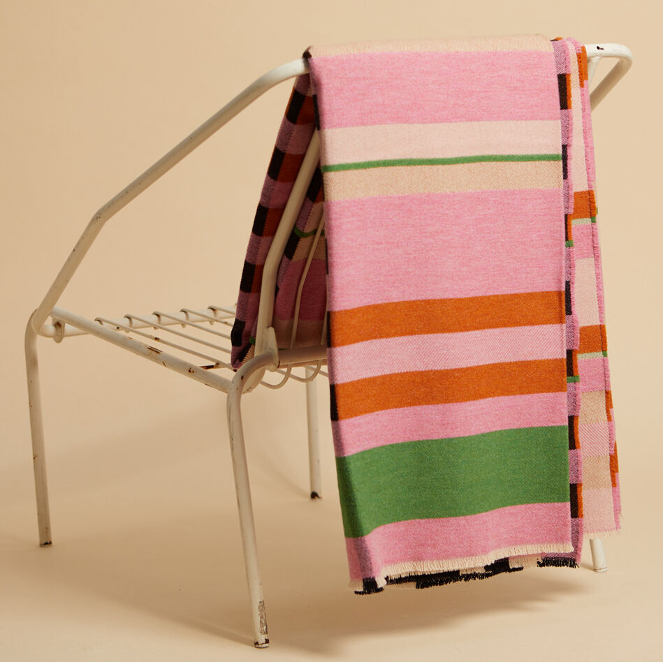 Eine Decke mit geometrischen Mustern in Rosa, Grün, Orange, Beige und Weiß liegt über einem minimalistischen weißen Stuhl vor einem neutralen Hintergrund; Bauhaus Stil.