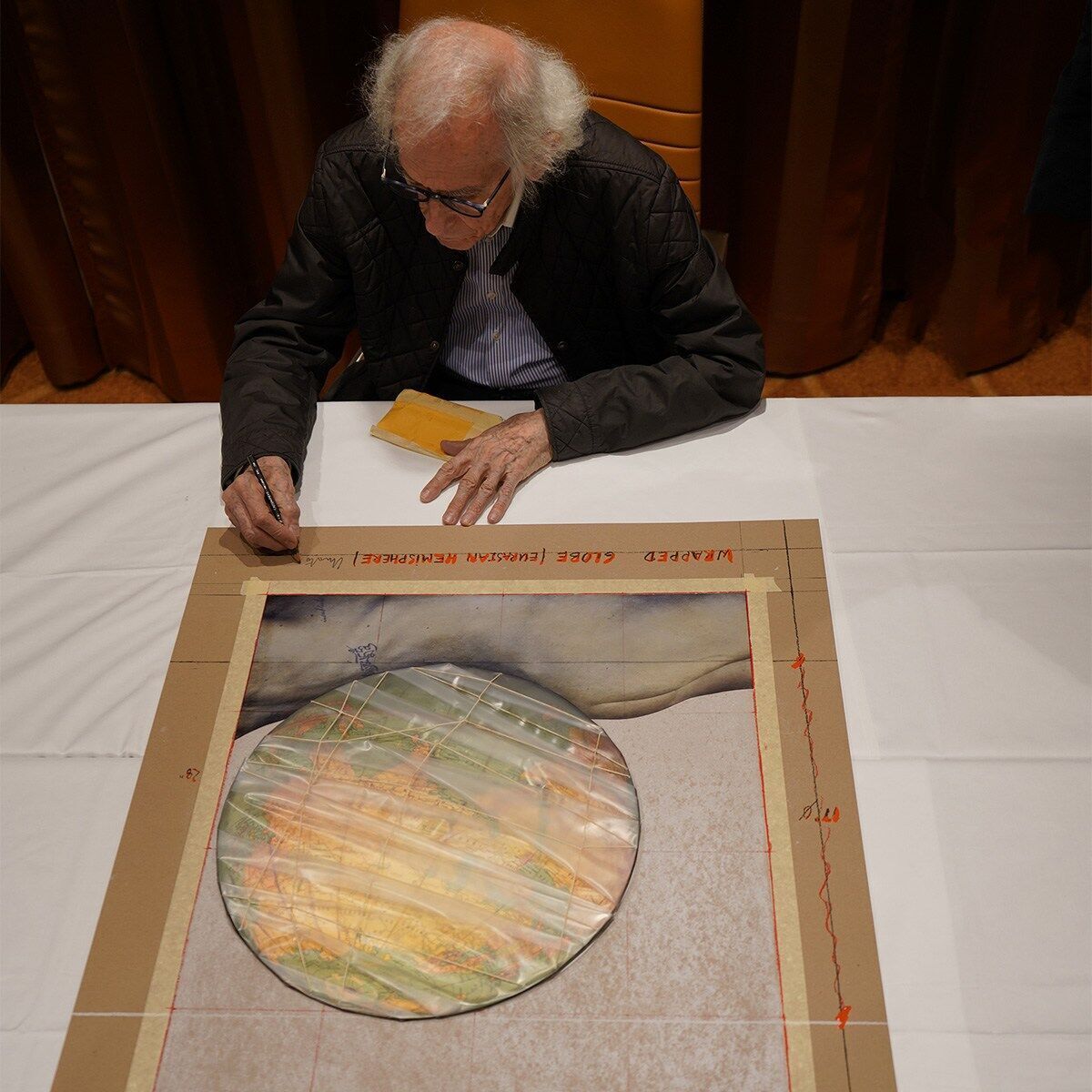 Ein älterer Mann mit einer Brille sitzt an einem Tisch und signiert ein Kunstwerk mit einem runden, in Stoff gewickelten Globus. Das Kunstwerk ist auf einer flachen Oberfläche platziert und von einem rechteckigen Rahmen umgeben.