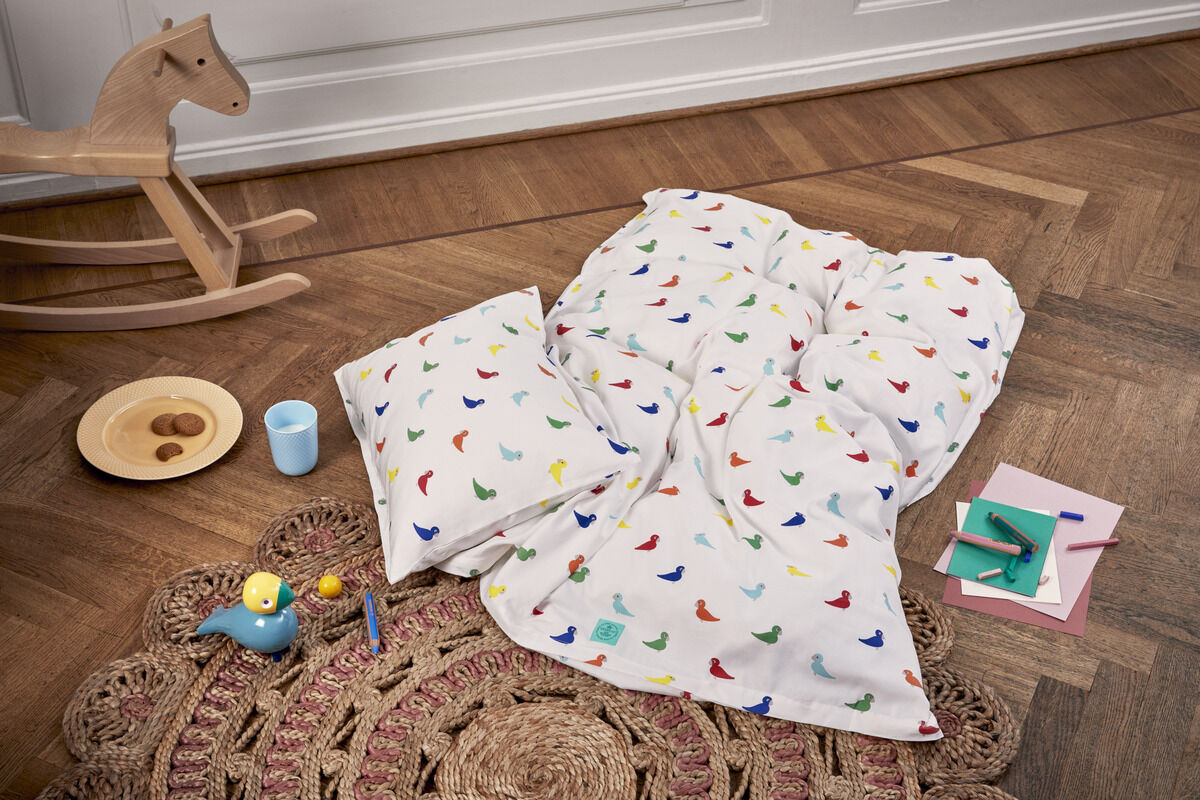 Singvogel Sunshine von Kay Bojesen ist auf einer farbenfrohen Kinderbettwäsche mit Vogelmotiven abgebildet, zusammen mit Spielzeug auf einem Holzboden. Das Szenario beinhaltet zudem eine Schaukelpferd und eine handgefertigte Unterlage.