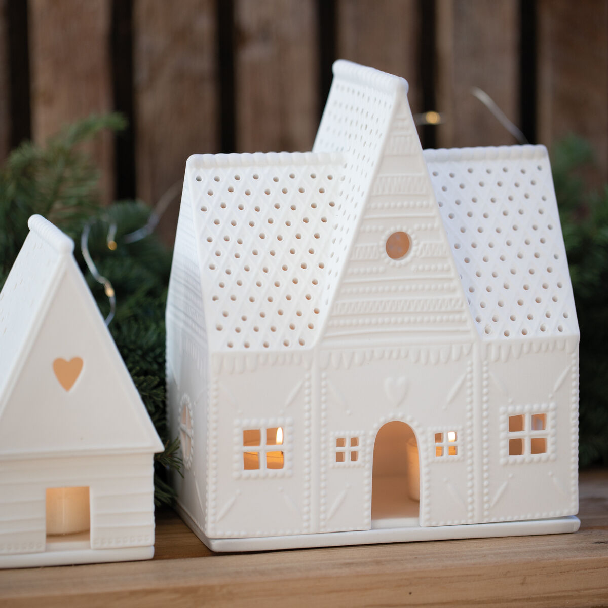 Ein keramisches Lichthaus in Form eines Lebkuchenhauses, das auf einem Holztisch steht. Das Haus hat filigrane Details und strahlt warmes Licht durch kleine Fenster und Öffnungen. Im Hintergrund sind grüne Nadelzweige zu sehen.