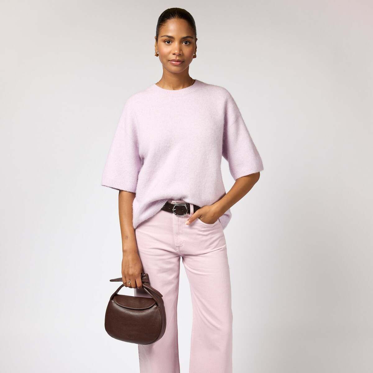 Eine Frau steht vor einem hellen Hintergrund und trägt ein hellrosa Oberteil und eine hellrosa Hose. Sie hält eine dunkelbraune Handtasche im Croissant-Stil in einer Hand und trägt spitze braune Schuhe. Das Outfit wirkt elegant und modern.