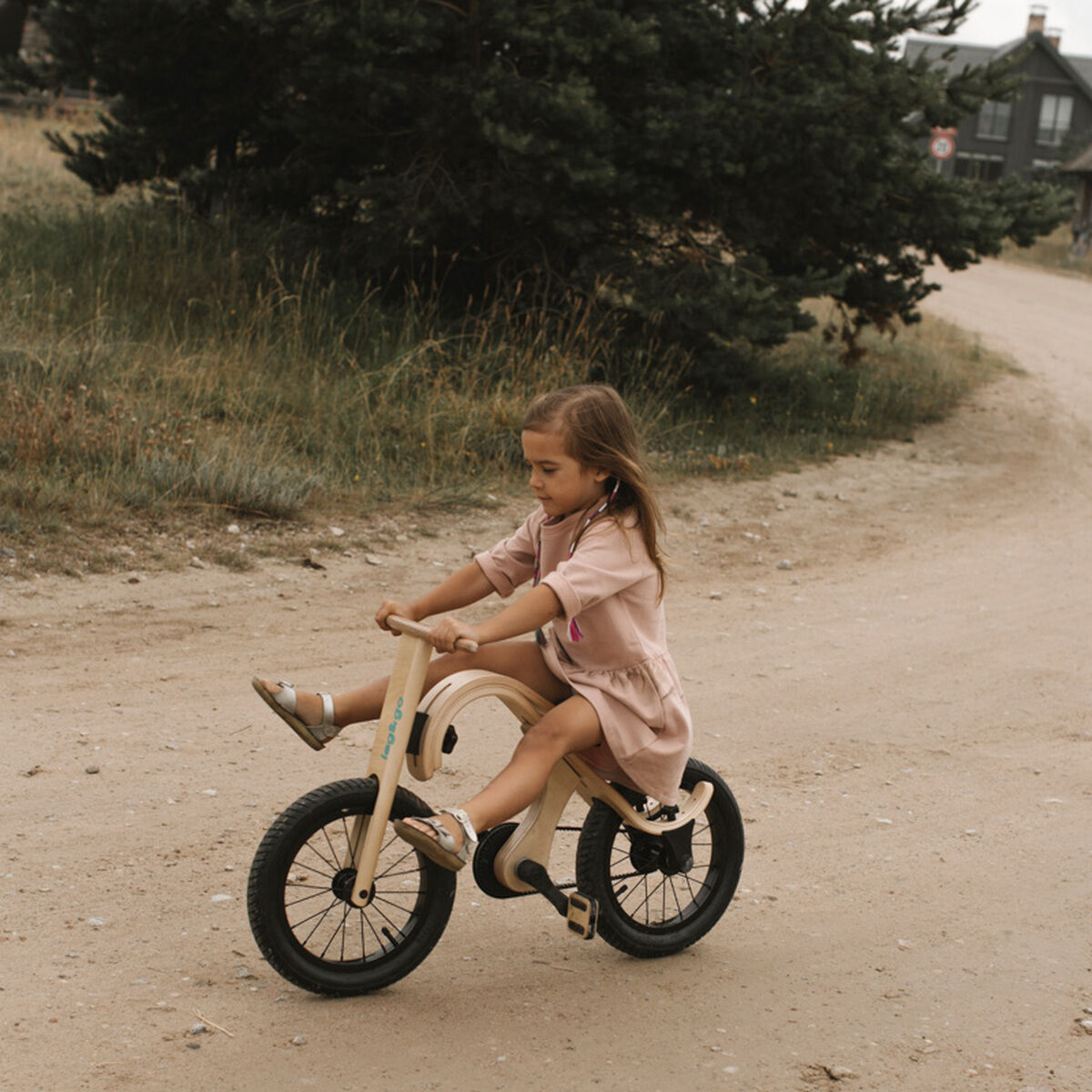 Ein Kind fährt draußen auf einem Balance Board mit montierten Pedalen über einen sandigen Weg. Das Board hat ein natürliches Holzdesign mit schwarzen Reifen. Im Hintergrund sind Bäume und ein Gebäude zu sehen.
