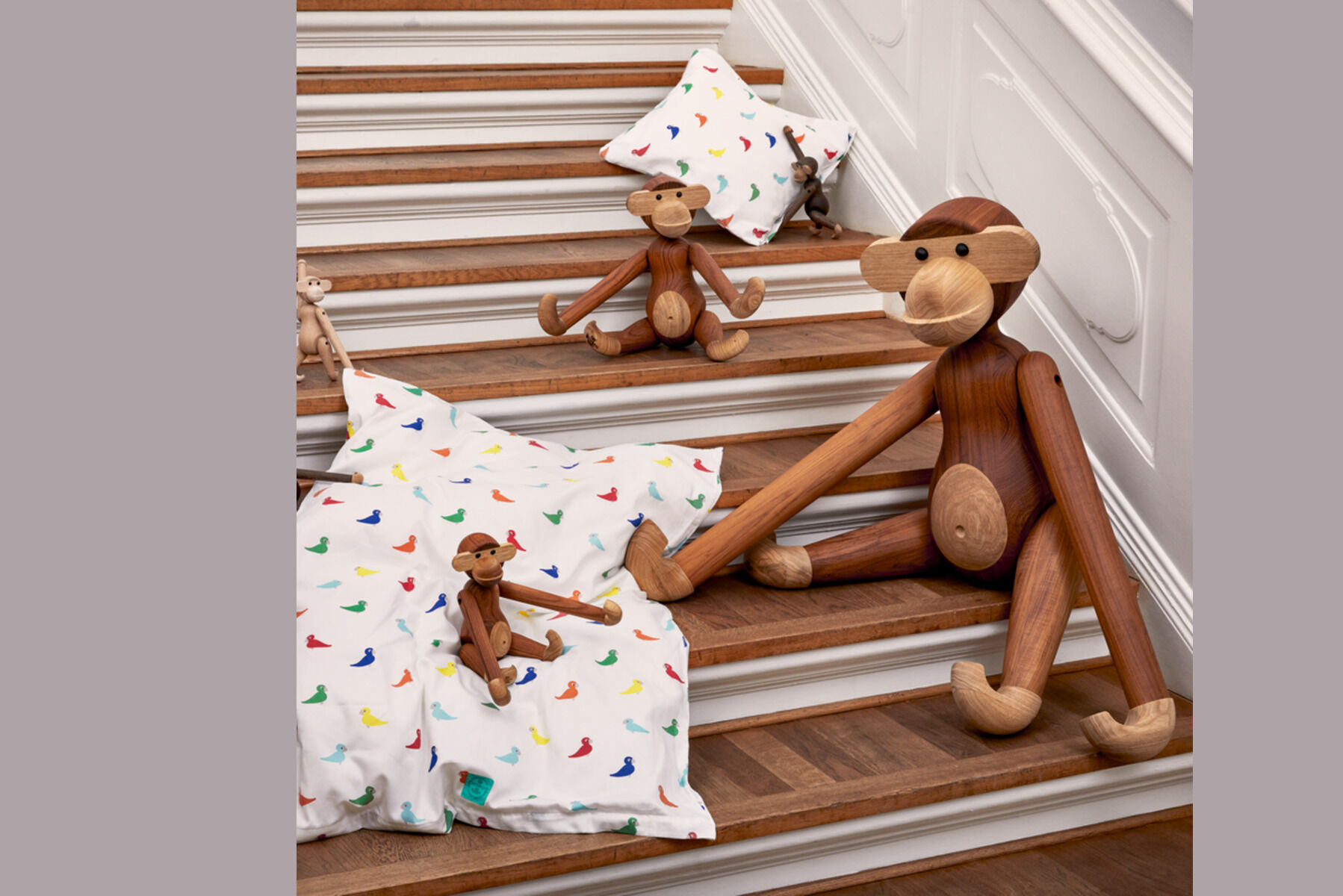 Mehrere Holzfiguren in Form von Affen sitzen auf einer Treppe. Die Figuren sind aus braunem Holz und befinden sich neben weißen Kissen mit bunten Mustern.