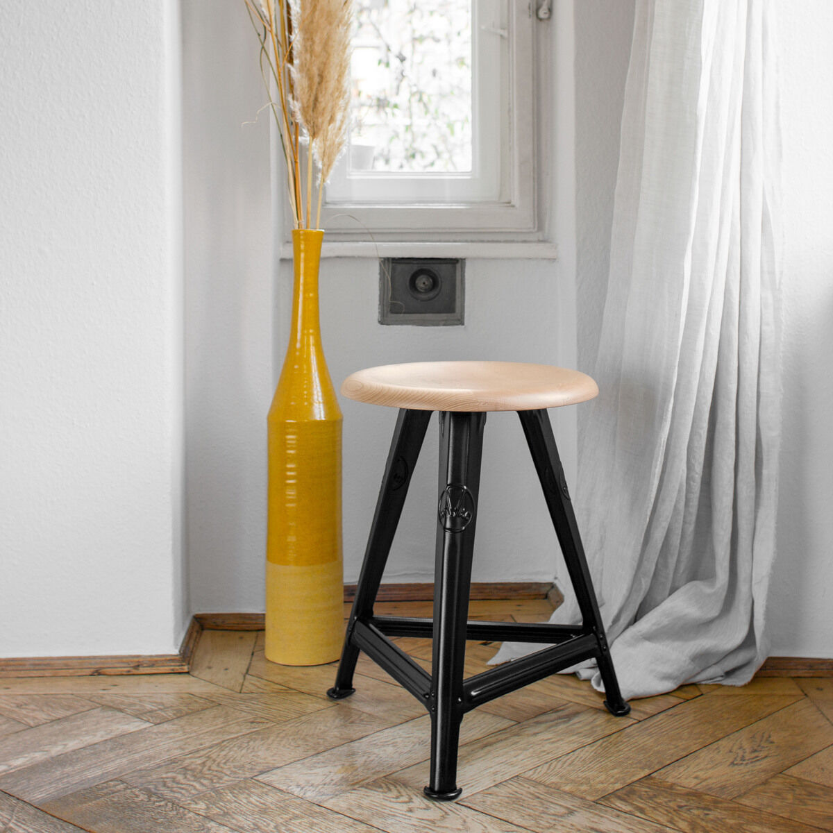 Ein moderner Hocker, genannt Rowac-Schemel »Eiche Mattschwarz«, ist in einem gemütlichen Raum platziert. Er hat eine runde Sitzfläche aus Eichenholz und vier mattschwarze Metallbeine. Der Hocker steht auf einem hölzernen Boden, und neben ihm befindet sich eine hohe gelbe Vase mit Trockenblumen. Im Hintergrund hängen weiße Vorhänge um ein Fenster.