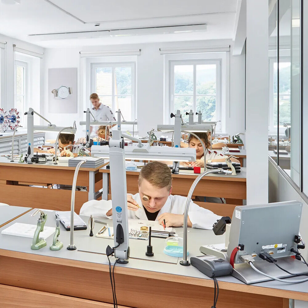 Lichtdurchflutete Uhrmacherwerkstatt bei NOMOS Glashütte: Mehrere Uhrmacher arbeiten konzentriert an Holztischen, ausgestattet mit Lupen, Präzisionswerkzeugen und modernen Lampen. Im Hintergrund große Fenster, klare Linien und ruhiges, modernes Design.