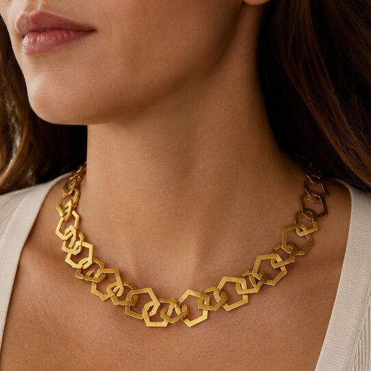 Detailaufnahme einer Frau, die eine massive goldfarbene Halskette aus vergoldetem Messing trägt. Die Kette besteht aus ineinandergreifenden geometrischen Elementen. Das Produkt ist das Schmuckset »Rosa« von Ars mundi, elegant und auffällig designt.