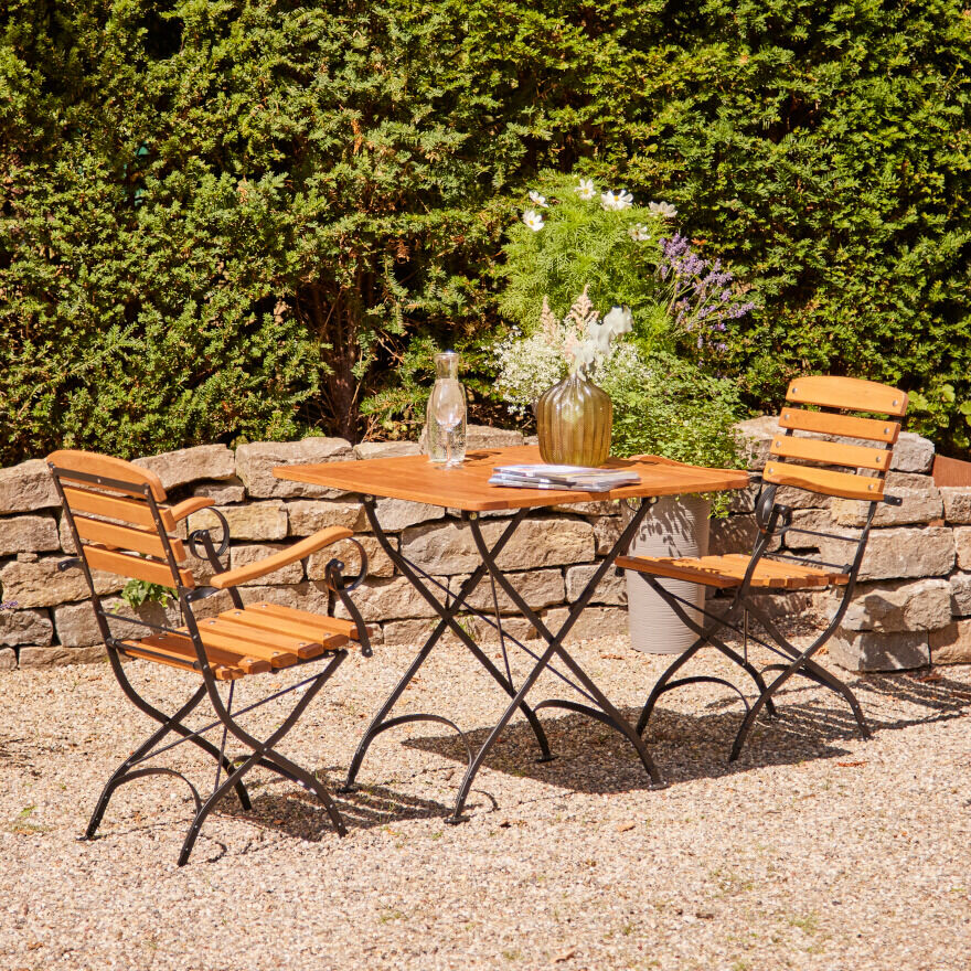 Der Klapptisch 'Peru III' mit den Maßen 80x80 cm steht im Garten auf einem Kiesboden. Er besteht aus Holzlatten mit einem schwarzen Metallrahmen. Auf dem Tisch stehen eine Glasvase mit Blumen und eine Karaffe. Im Hintergrund sind eine grüne Hecke und ein Steinwall zu sehen.