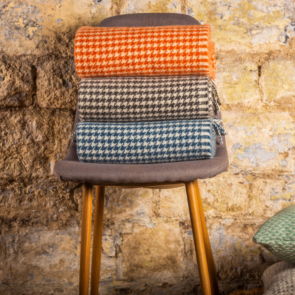 Drei in Hahnentrittmuster gestaltete Decken in den Farben Orange, Grau und Blau sind auf einem grauen Stuhl gestapelt. Eine grün-beige Decke liegt daneben auf einem passenden Kissen vor einer beigen Steinwand.