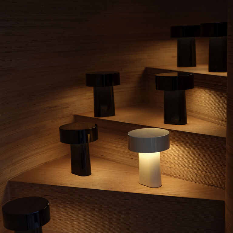 Mehrere tragbare Lampen des Modells 'Oblong' stehen auf den Stufen einer Holztreppe. Eine der Lampen leuchtet mit warmweißem Licht, während die anderen ausgeschaltet sind. Das Design ist modern und minimalistisch. Die Szene strahlt eine gemütliche und stilvolle Atmosphäre aus.