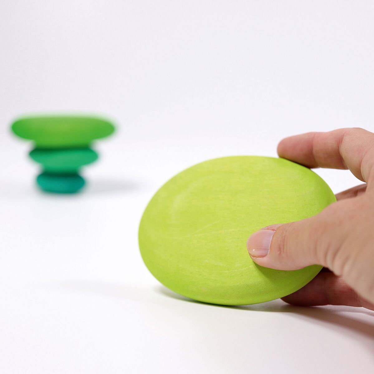 Detailreiches Foto zeigt eine Kinderhand, die einen ovalen, hellgrünen, flach geschliffenen Holzstein von GRIMM’S hält. Im unscharfen Hintergrund sind weitere, übereinandergestapelte grüne Holzsteine zu sehen. Der Hintergrund ist weiß, die Szene minimalistisch gestaltet.