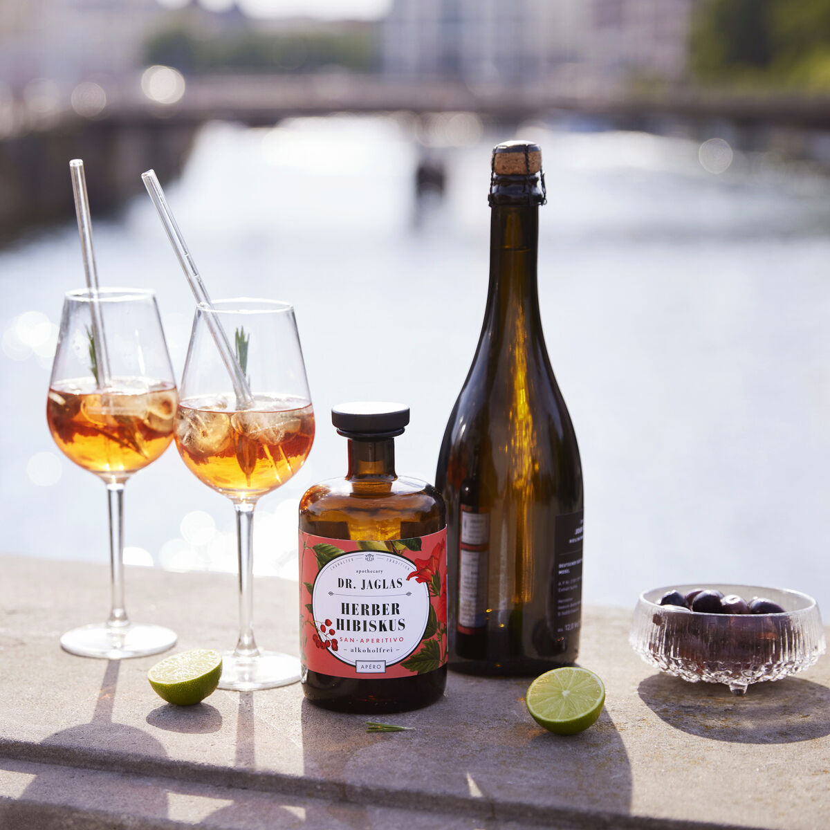 Dieses Bild zeigt eine Flasche 'Herber Hibiskus' alkoholfreier Aperitif, eine geöffnete Sektflasche, zwei gefüllte Weingläser mit Strohhalmen und einige Limettenscheiben, die auf einem steinernen Geländer an einem Flussufer arrangiert sind. Im Hintergrund sieht man den Fluss und eine verschwommene, sonnige Landschaft.