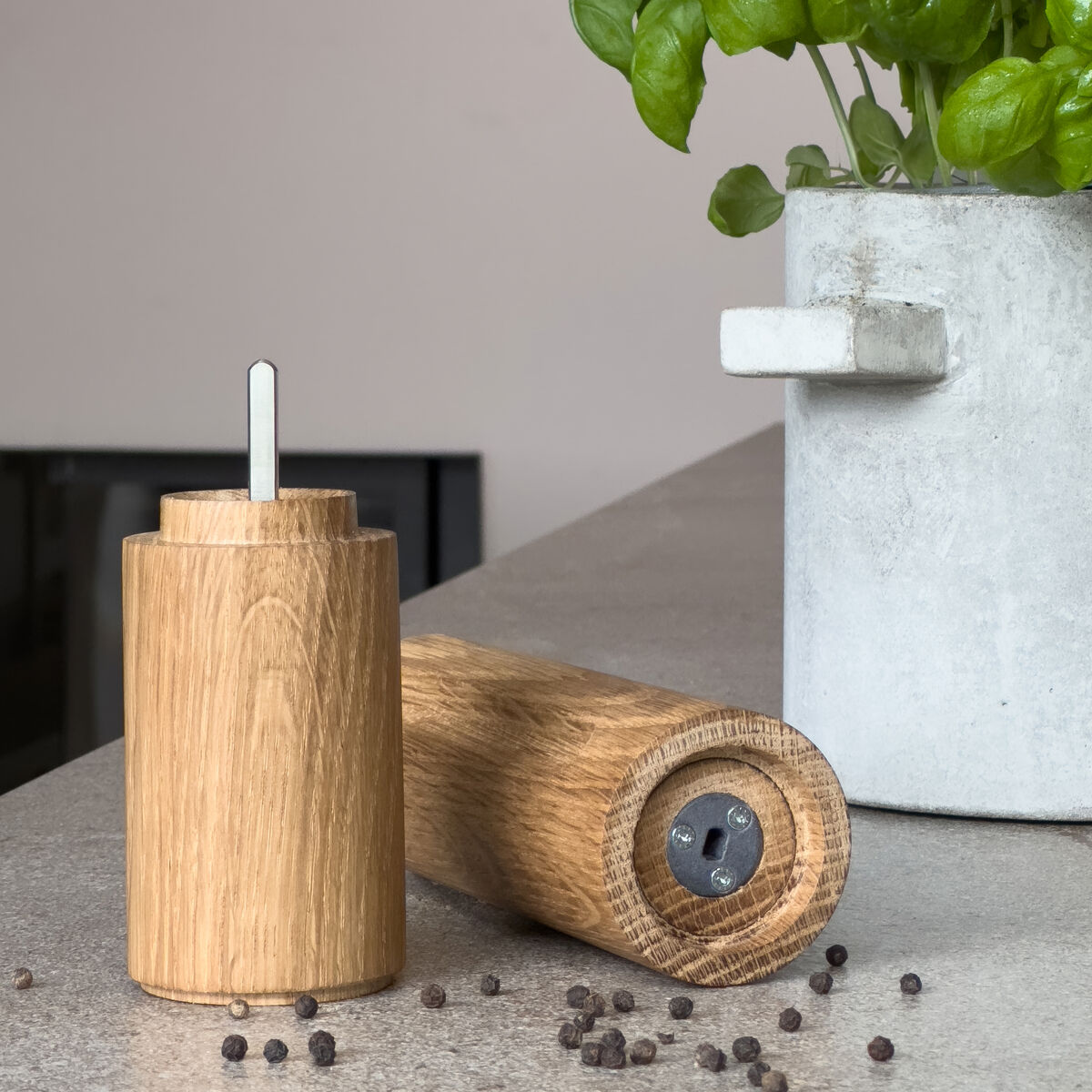 Zwei Gewürzmühlen vom Typ Gewürzmühle »TURN« aus massiv geölter Eiche der Marke finbeaux, Modell von Christoph Schoen, stehen auf einer Küchenoberfläche neben einem Topf mit grünem Basilikum. Einige Pfefferkörner sind verstreut. Dezente, moderne Szene.