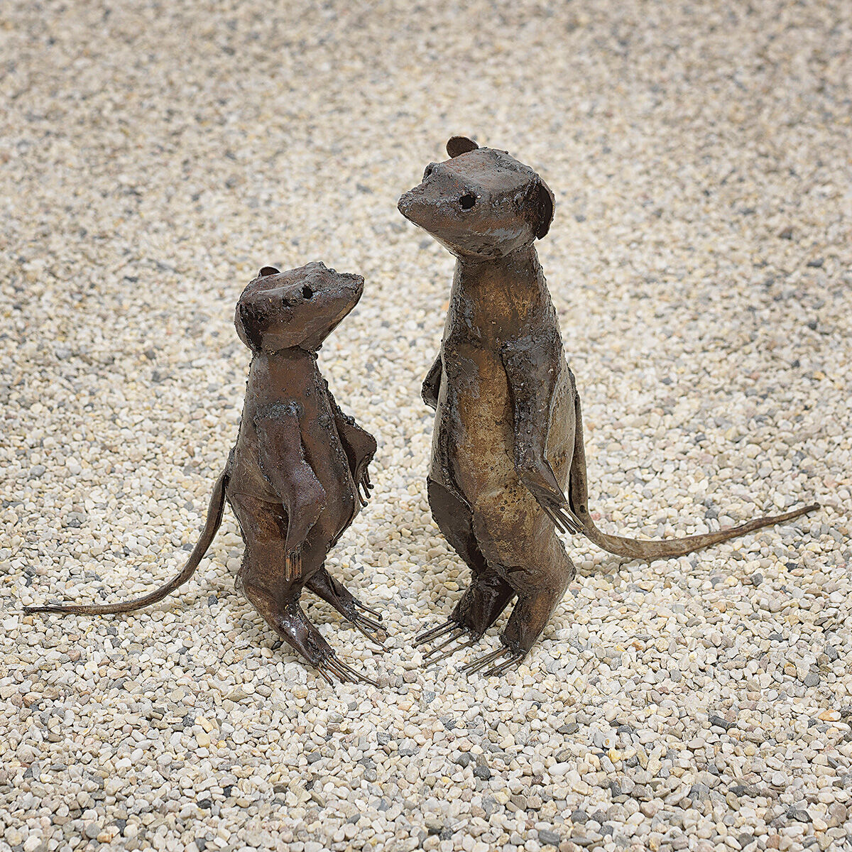 Zwei Gartenfiguren aus Metall in rostbrauner Optik, ein großes und ein kleines Erdmännchen, stehen auf einem kiesbedeckten Boden.