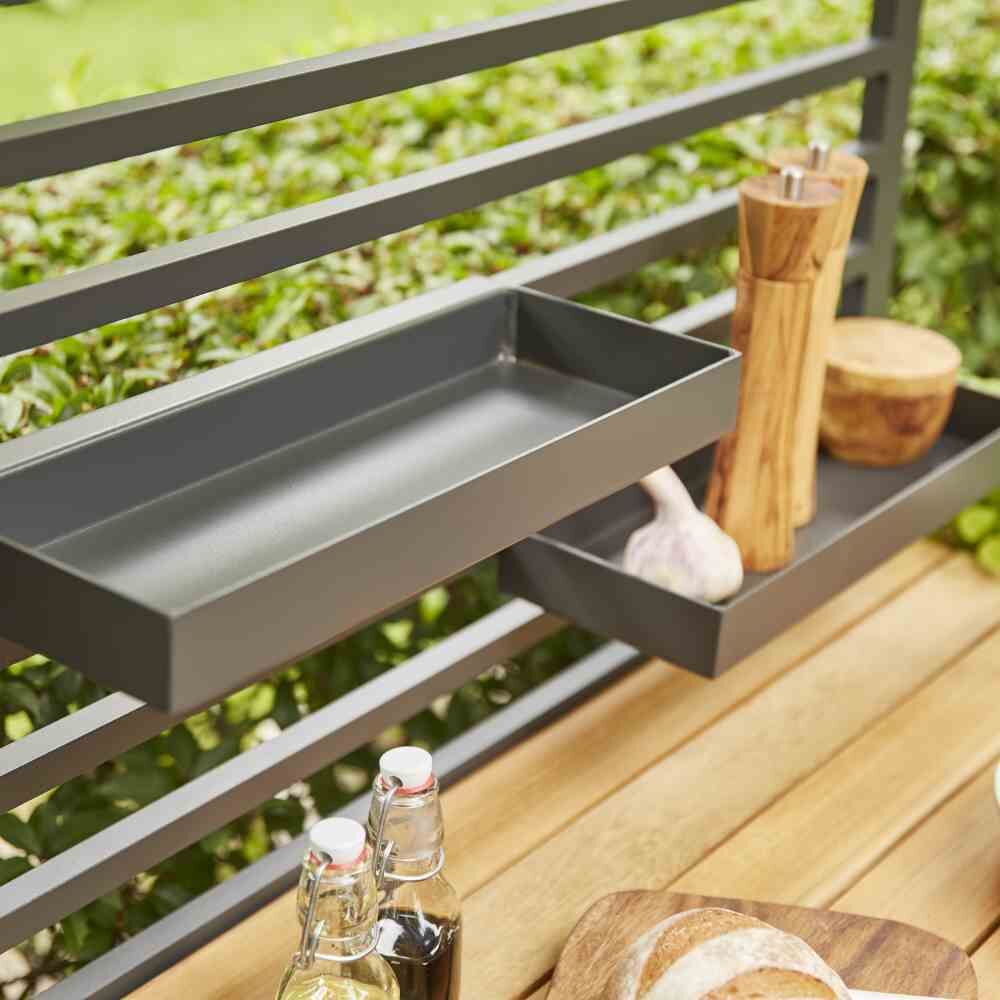 Ein modernes Outdoor-Küchenmodul mit Metallrahmen, einem Tablett und Flaschen darauf, sowie Gartenhintergrund mit Sichtschutz.