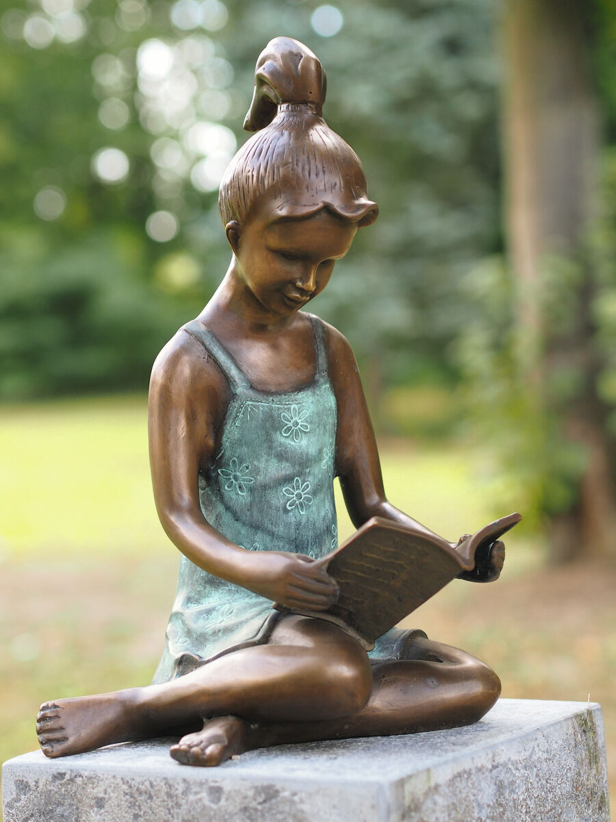 Bronzeskulptur eines lesenden Mädchens sitzend auf einem Stein im Garten, umgeben von Grünpflanzen.