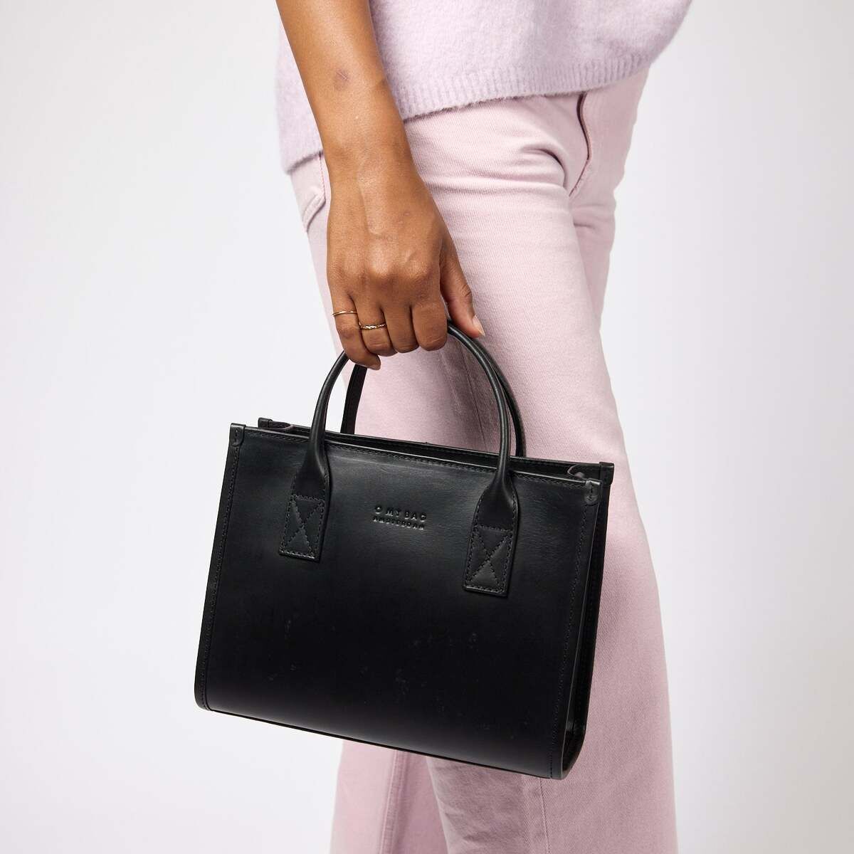Eine Person trägt eine schwarze, mittlere Handtasche »Jackie Midi« mit kurzem Tragegriff. Die Person ist in hellrosa Hose und Oberteil gekleidet. Die Tasche wirkt schlicht und modern und passt zu einem eleganten, minimalistischen Stil.