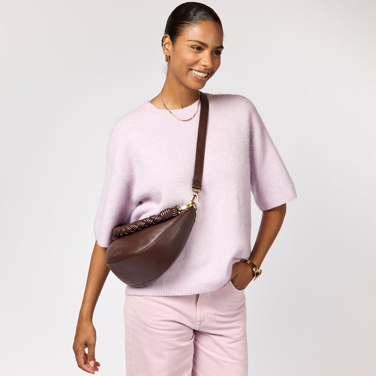 Frau mit dunkelbrauner Crossbody-Ledertasche „Leo“ von O My Bag mit dekorativ geflochtenem Griff, trägt einen fliederfarbenen, kurzärmligen Pullover und rosa Hose vor einfarbig hellem Hintergrund. Die Tasche wirkt edel, weich und ist elegant am Oberkörper getragen.