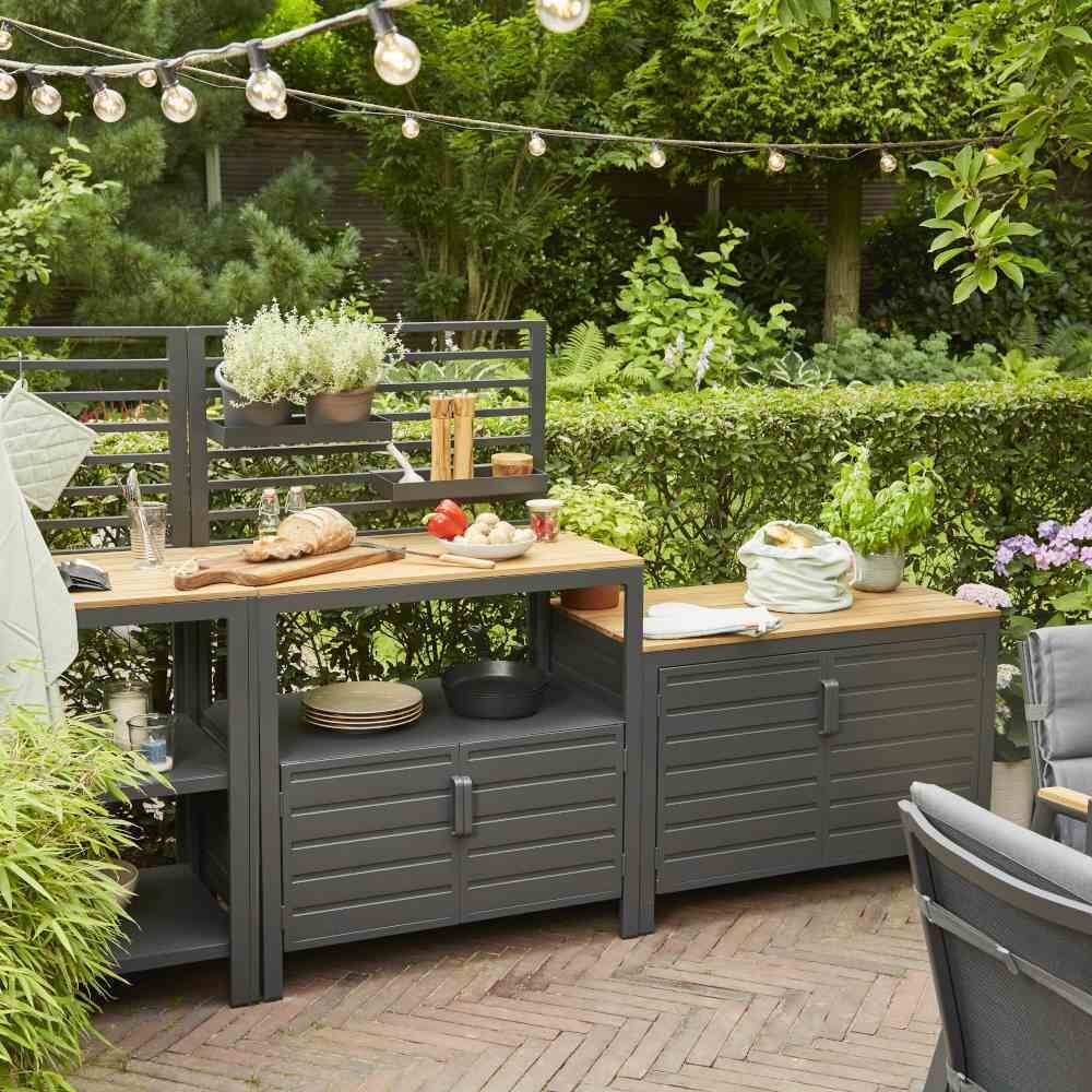 Ein 'Outdoor Kitchen Mittelteil niedrig »Sierra«' in einem Garten, bestehend aus einem grauen Holztresen mit Regalen und Schubladen. Darauf stehen Küchenutensilien und Pflanzen. Im Hintergrund dichter grüner Bewuchs.