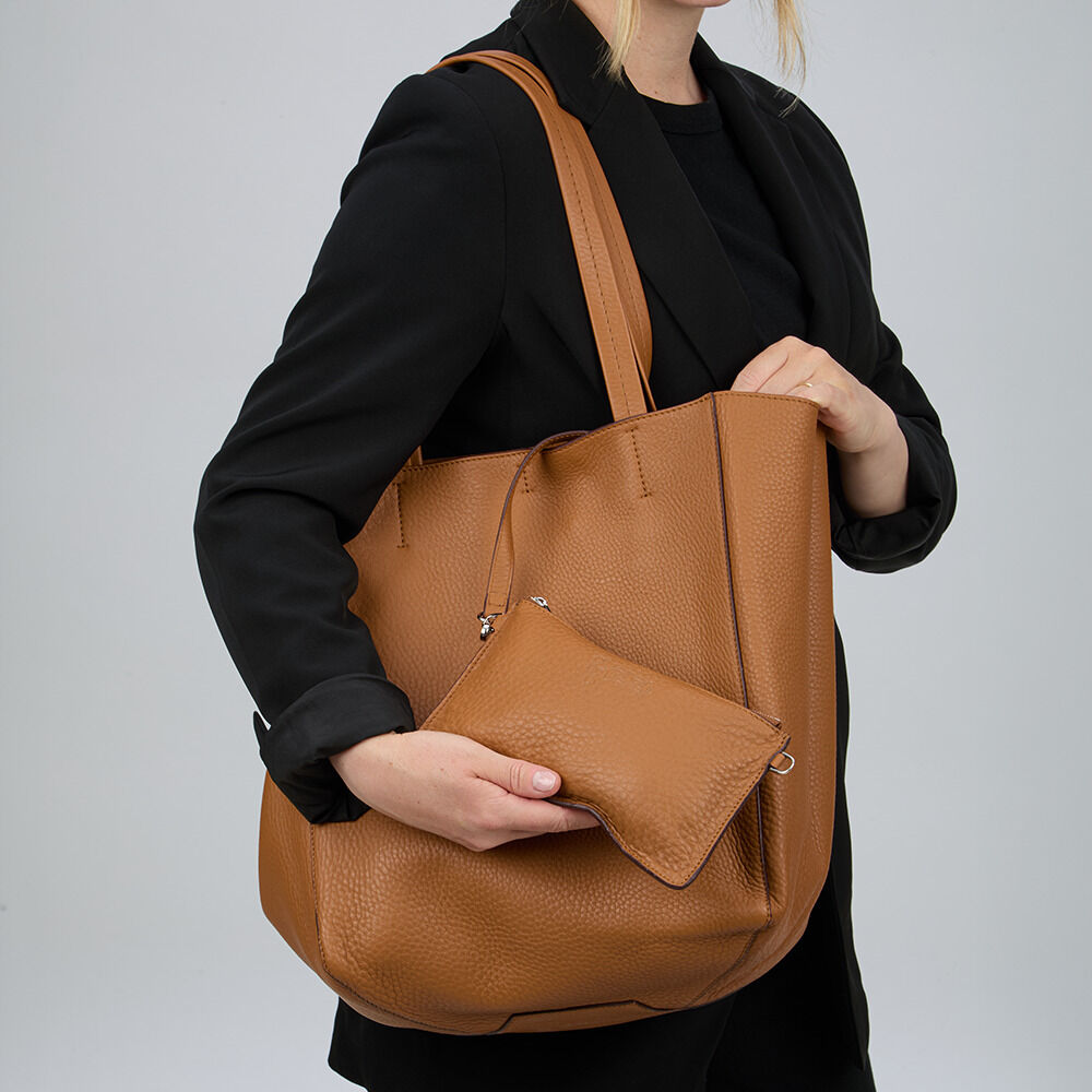 Frau in schwarzer Kleidung hält eine hochwertige cognacfarbene Ledertasche von Guerer & Guerer. Die Tasche besitzt eine feine Narbung, längere Henkel sowie eine passende kleine Innentasche, die als Kosmetiktäschchen oder Geldbörse genutzt werden kann.