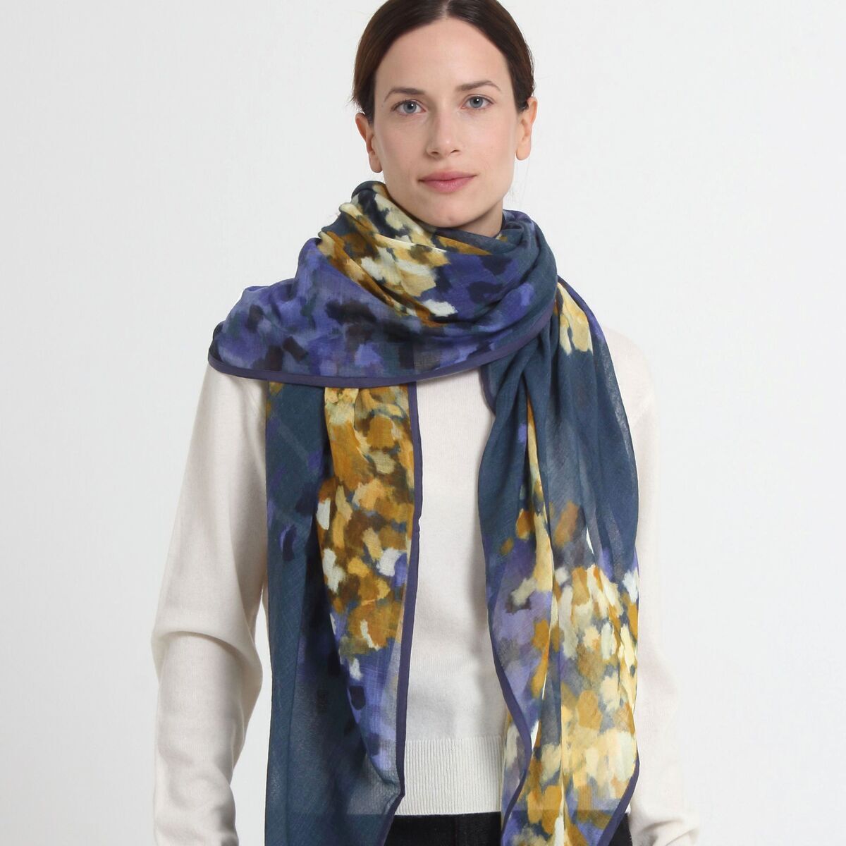 Dame trägt einen luxuriösen Wollschal »Hazel« von Shalls Barcelona. Der Schal besteht zu 100 % aus Schurwolle und zeigt ein abstraktes, florales Muster in Blau, Violett, Gelb und Weiß. Das Accessoire wirkt elegant und leger zugleich, ideal für Modebewusste.