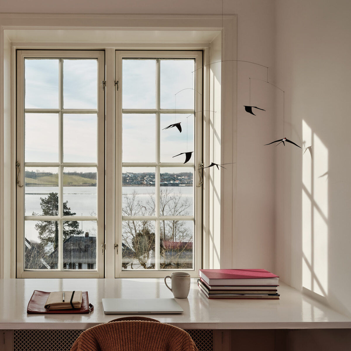 Ein modernes Mobile mit fliegenden Schwalben hängt vor einem Fenster mit Gittersprossen. Darunter steht ein Schreibtisch mit Büchern, einer Tasse und einem Notizbuch. Der Raum ist hell und lichtdurchflutet mit schlichter, minimalistischer Einrichtung.