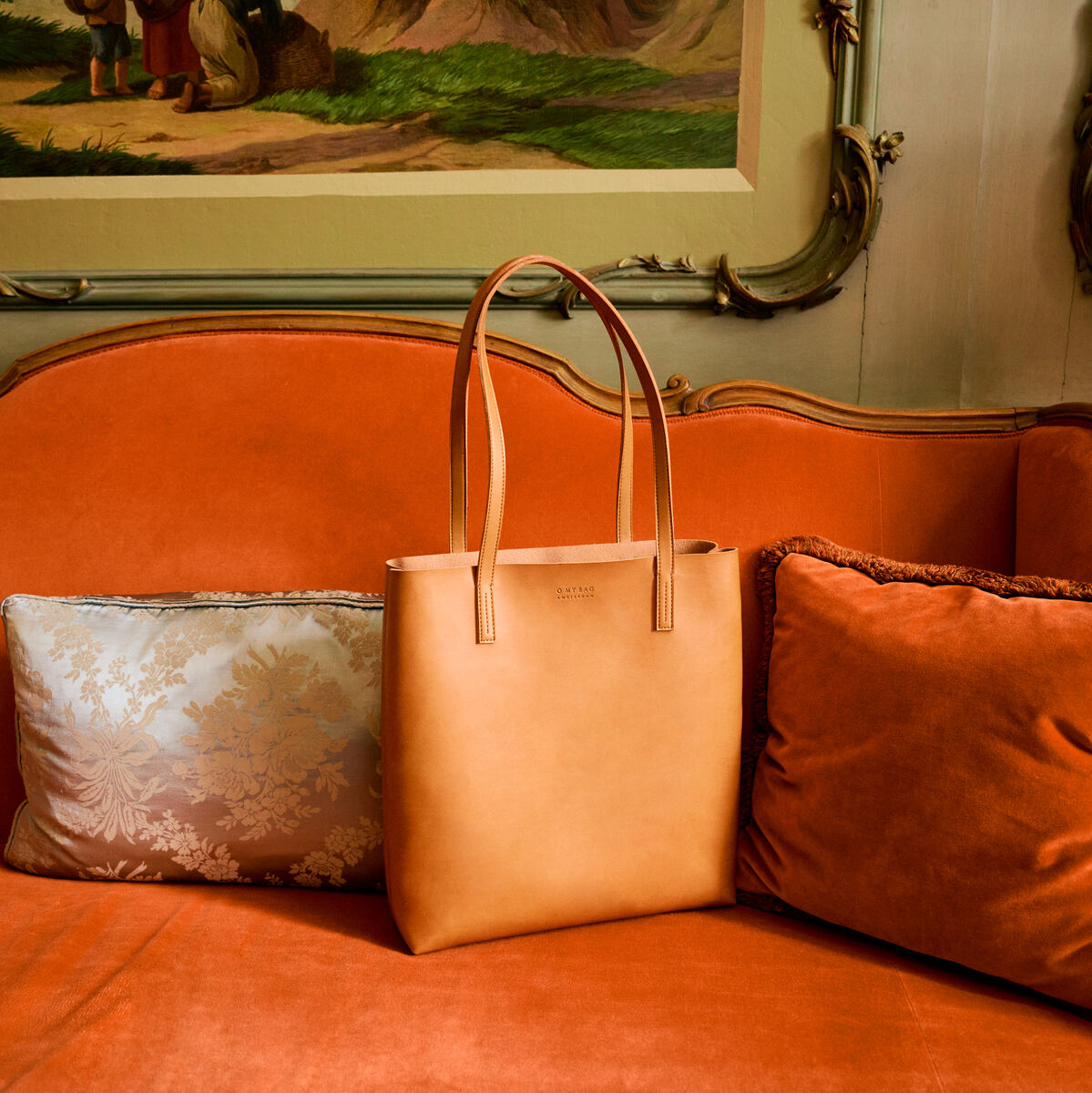 Beigefarbene Shopper-Tasche 'Georgia' aus Leder auf einem eleganten, orangefarbenen Sofa mit dekorativen Kissen im Hintergrund, in einem stilvollen Wohnraum.