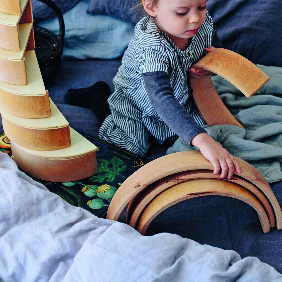 Ein Kind spielt mit einem Regenbogen-Holzspielzeug auf einem Bett. Das Spielzeug besteht aus mehreren halbkreisförmigen Holzteilen, die zusammen einen Regenbogen bilden. Das Kind greift eines der Holzteile und ist in vertieftes Spiel vertieft. Im Hintergrund sind Decken und Kissen zu sehen. Das Holzspielzeug fördert die Kreativität und Motorik der Kinder.