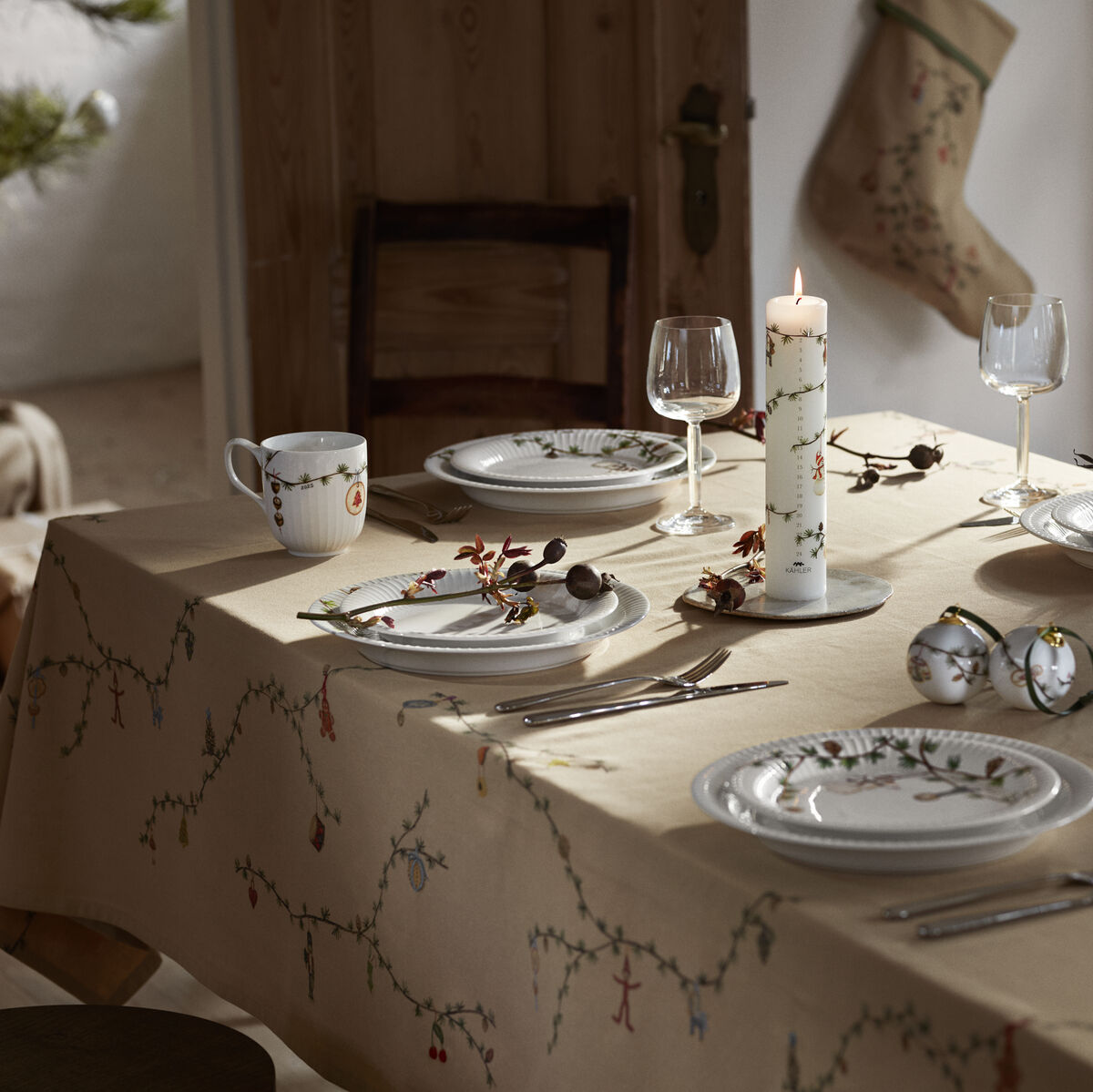 Advents­kerze »Hammershøi« 2025 steht angezündet und zentral auf einem festlich gedeckten Tisch mit winterlich-weihnachtlicher Tischdecke. Umgeben von Porzellantellern mit filigranen Motiven, Gläsern, Keramikbechern und kleinen Dekorationselementen, verbreitet die Szene gemütliche Weihnachtsstimmung in hellem, einladendem Tageslicht.