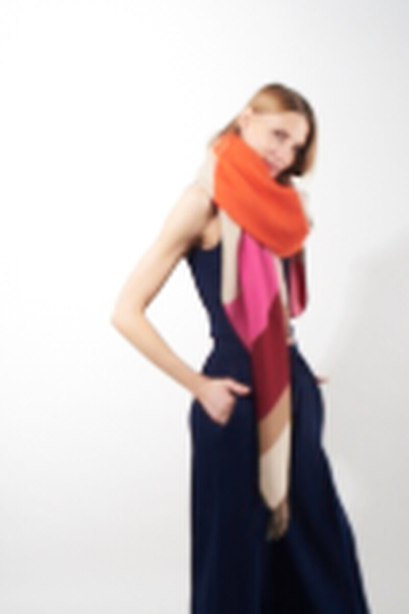Frau trägt einen farbenfrohen Schal aus Alpaka-Wolle in Rot-, Orange-, Beige- und Rosatönen, kombiniert mit einem dunkelblauen ärmellosen Kleid. Hintergrund ist weiß.