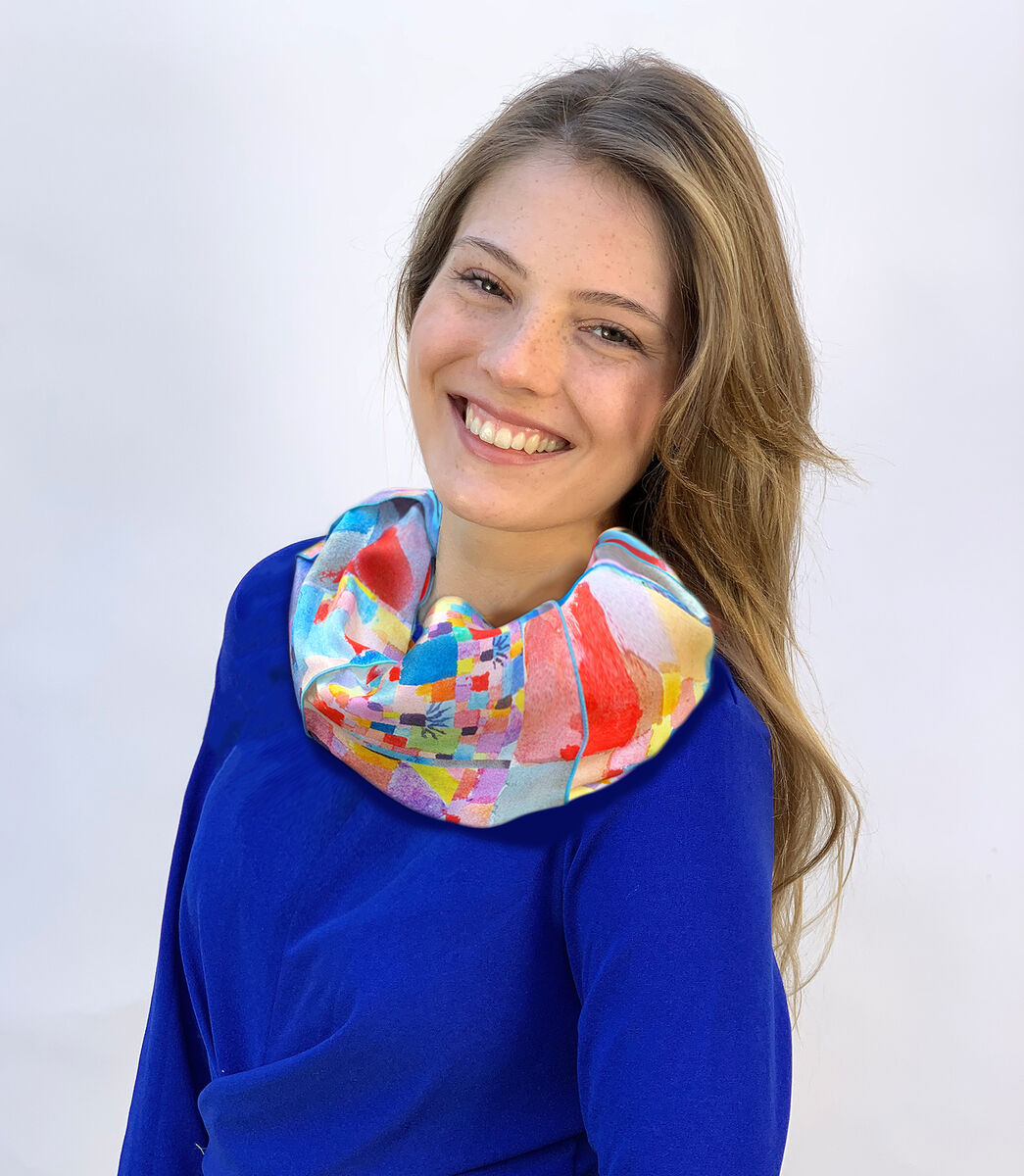 Eine lächelnde Frau trägt einen Loopschal mit dem Design 'Südliche Gärten' von Paul Klee. Der Schal zeigt ein farbenfrohes und abstraktes Muster in Blau, Rot, Pink, Grün und Gelb. Die Frau trägt dazu ein blaues Kleid.