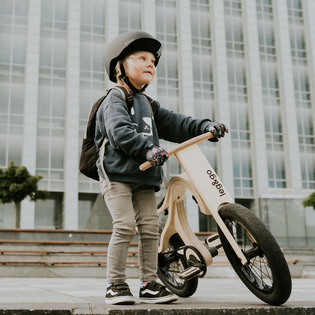 Ein Kind mit grauem Helm und Rucksack steht mit einem Balance-Board mit Pedalerweiterung auf einem urbanen Platz. Das Board besteht aus hellem Holz und die Pedale sind angesetzt. Im Hintergrund ein modernes Bürogebäude mit großen Fenstern und einigen grünen Bäumen.