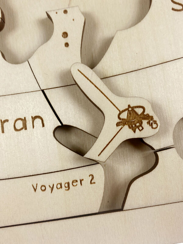 Nahaufnahme eines Holzpuzzles mit dem Motiv des Kosmos. Verschiedene Puzzleteile zeigen Planeten und Raumsonden. Eines der Puzzleteile ist mit der Aufschrift 'Voyager 2' beschriftet.