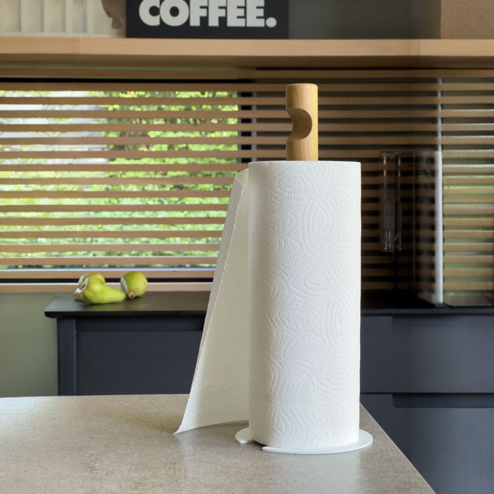 Küchenrollenhalter »GIVE« von finbeaux auf einer hellen Arbeitsplatte: Eine weiße Küchenpapierrolle steht auf einem flachen, runden weißen Sockel und wird von einem hellen Holzstab mit seitlicher Griffmulde gehalten. Ein Blatt hängt nach links herab. Im Hintergrund sind dunkle Küchenschränke, Holzjalousien, zwei grüne Birnen und ein Schild mit „COFFEE.“ zu sehen.