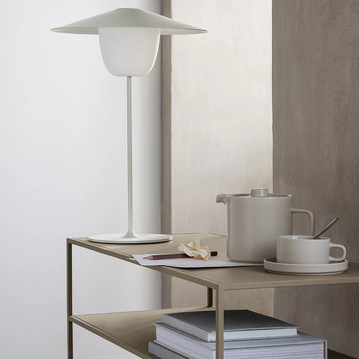 Mobile LED Tischleuchte »ANI« im modernen Design auf einem Regal mit Teekanne und Tasse. Minimalistisches Weiß und Grau dominieren die Farbpalette mit klaren Linien und schlichtem Äußeren.
