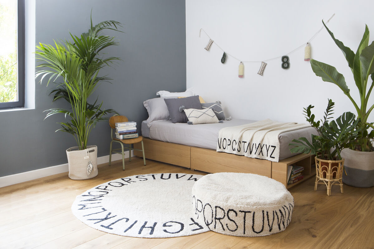 Modernes Kinderzimmer mit einem Sitzkissen »Pouf ABC« in Form eines runden Teppichs mit Alphabet-Muster, einer grauen Wand, einem Holzfußboden, Pflanzen und einer einfachen Dekoration.