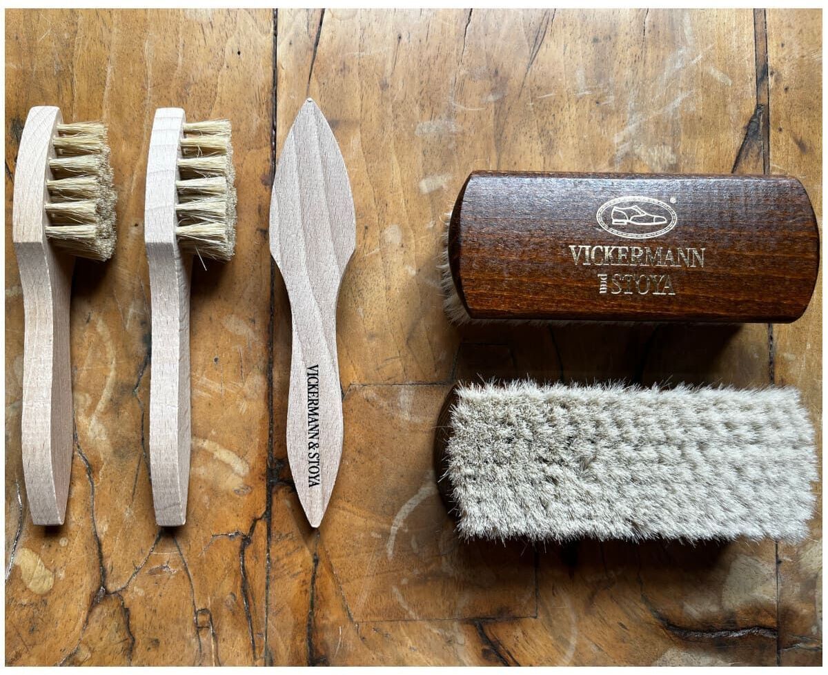 Lederpflege-Set bestehend aus zwei Holzbürsten mit Naturhaarborsten, einem hölzernen Pflegemesser und einer kleinen rechteckigen Holzbox mit dem Aufdruck 'Vickermann 8-SOH', präsentiert auf einem rustikalen Holztisch.