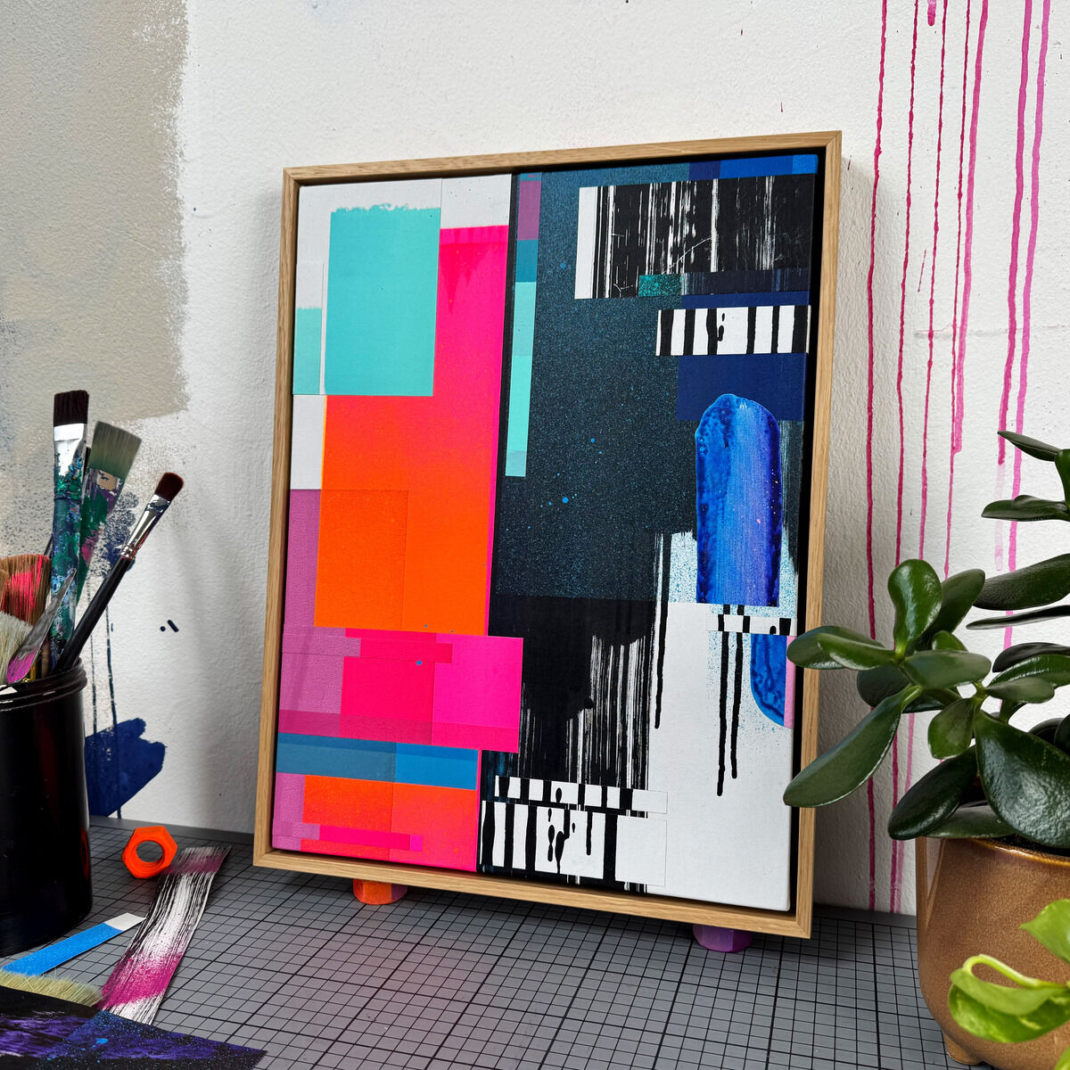 Handgefertigtes Unikat »Gratitude - No.10« von Daniel Lisson, Collage auf Baumwollgewebe im Eichenholzrahmen. Farbflächen in Pink, Türkis, Blau, Schwarz und Weiß, kombiniert mit linearen und gesprenkelten Strukturen. Umgeben von Malutensilien und modernen Akzenten.